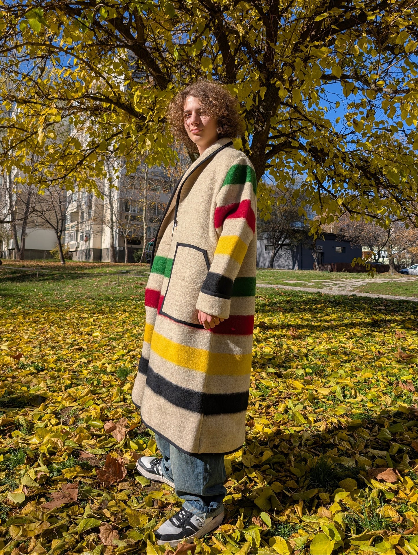 A young man wearing a long candy stripe robe coat, upcycled from a traditionally woven wool. The man is posing outside in a park.