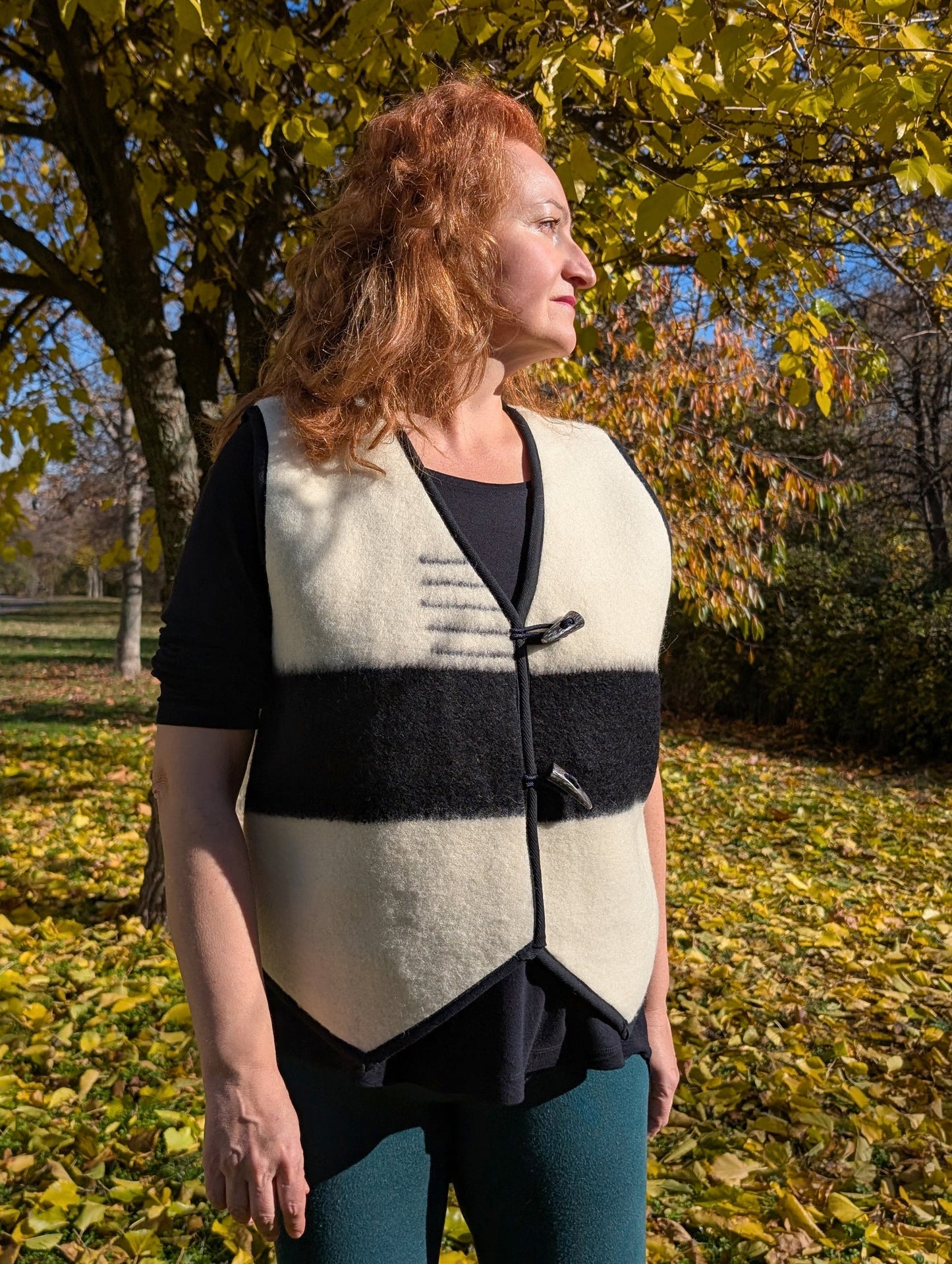 Woman wearing an off-white or ivory vest with a black statement stripe running across the front side. The vest is upcycled from a pure wool blanket and has black toggles.