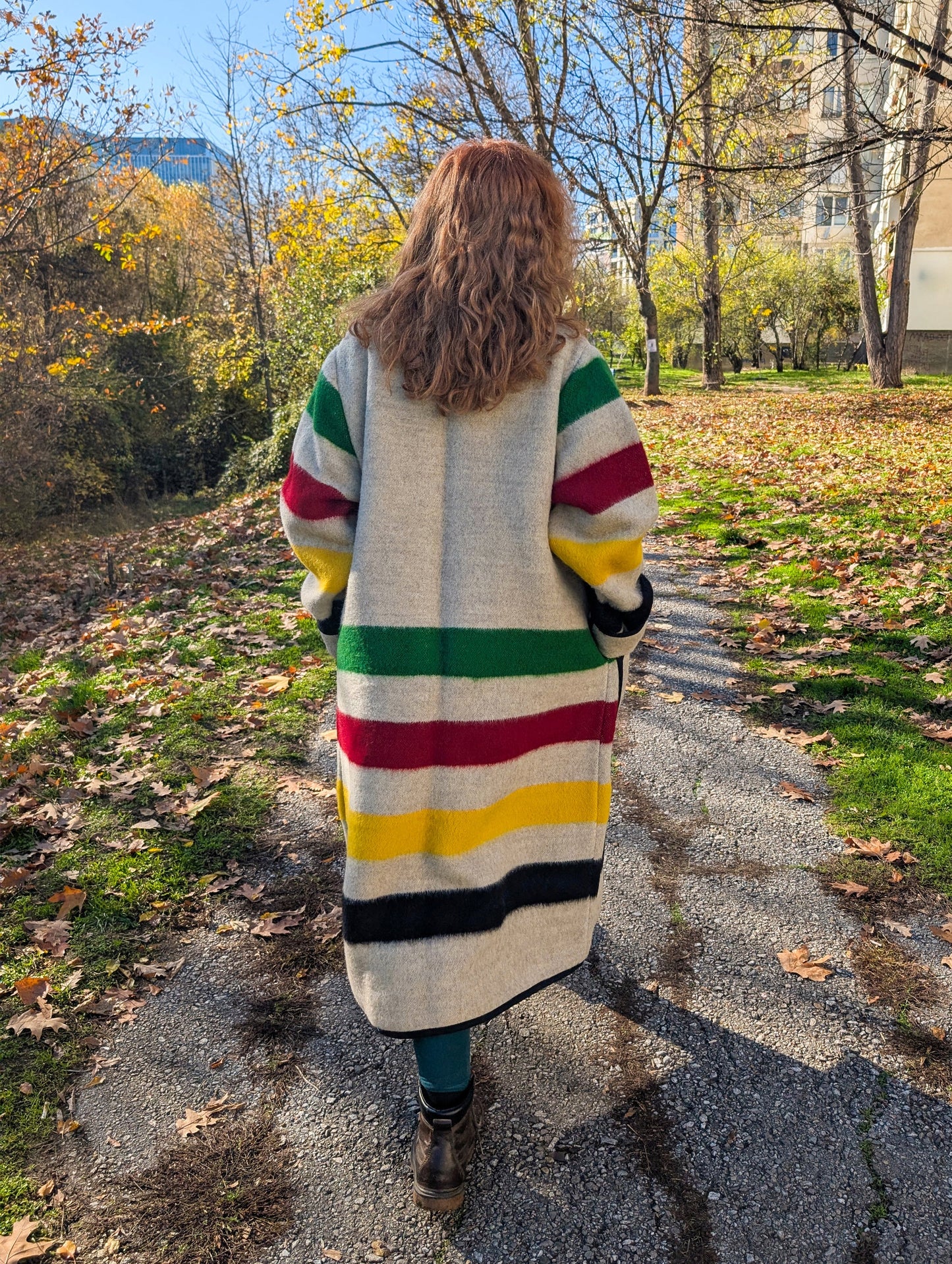 A woman posing a colorful striped long blanket jacket walking on a path with trees and buildings in the background. 