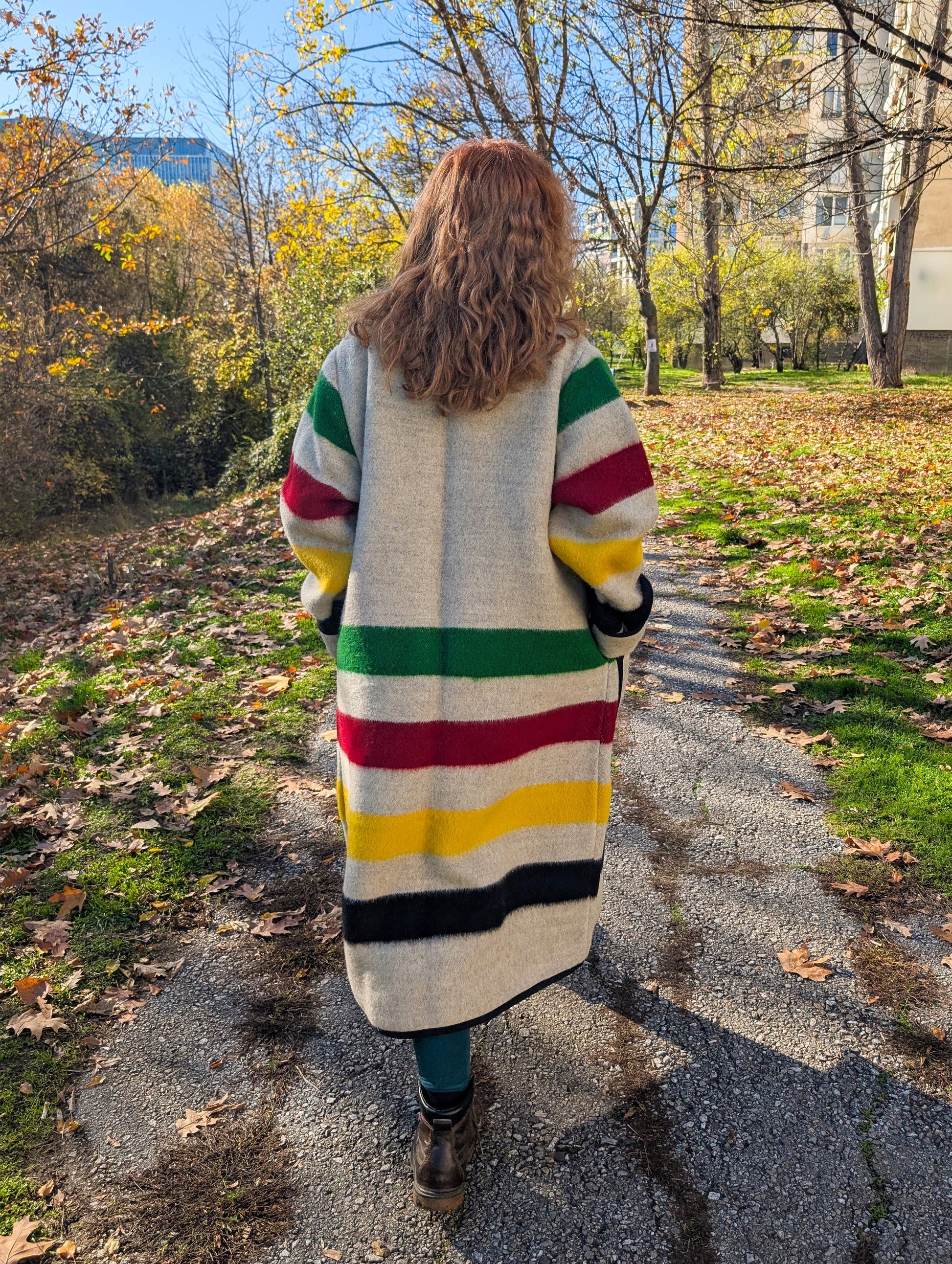 A woman posing a colorful striped long blanket jacket walking on a path with trees and buildings in the background. 