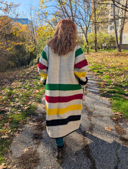 A woman posing a colorful striped long blanket jacket walking on a path with trees and buildings in the background. 