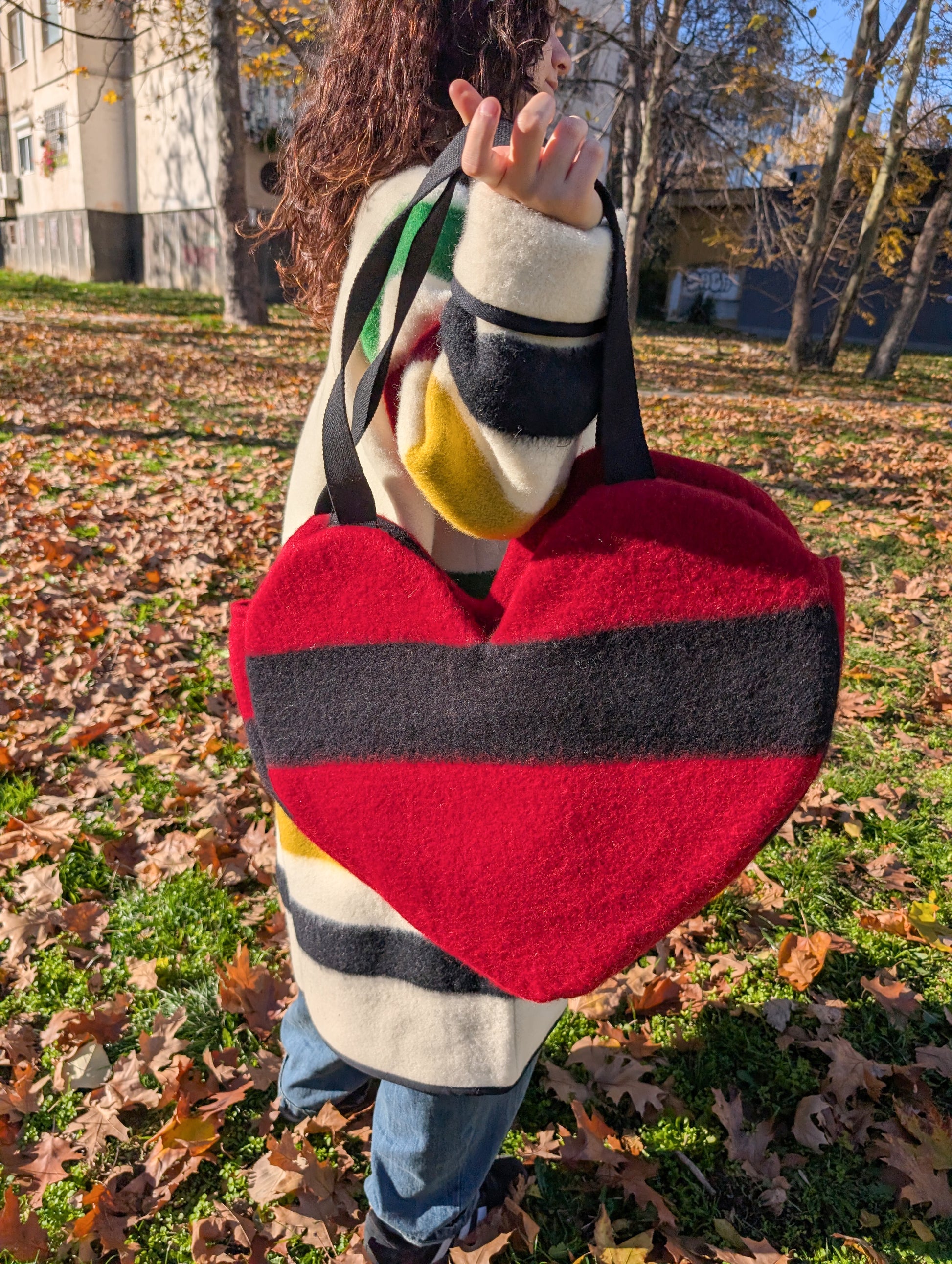 Woman holding a heart-shaped bag with a wide black stripe in an outdoor setting with fallen leaves.