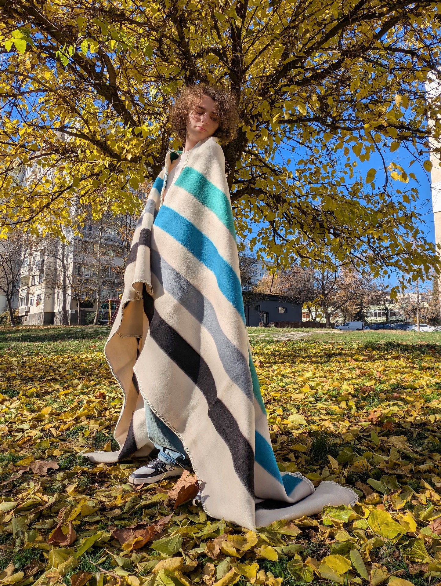 Person wrapped in a striped handspun wool blanket standing in a park with autumn leaves and trees.