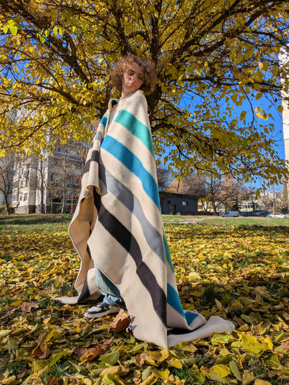 Person wrapped in a striped handspun wool blanket standing in a park with autumn leaves and trees.