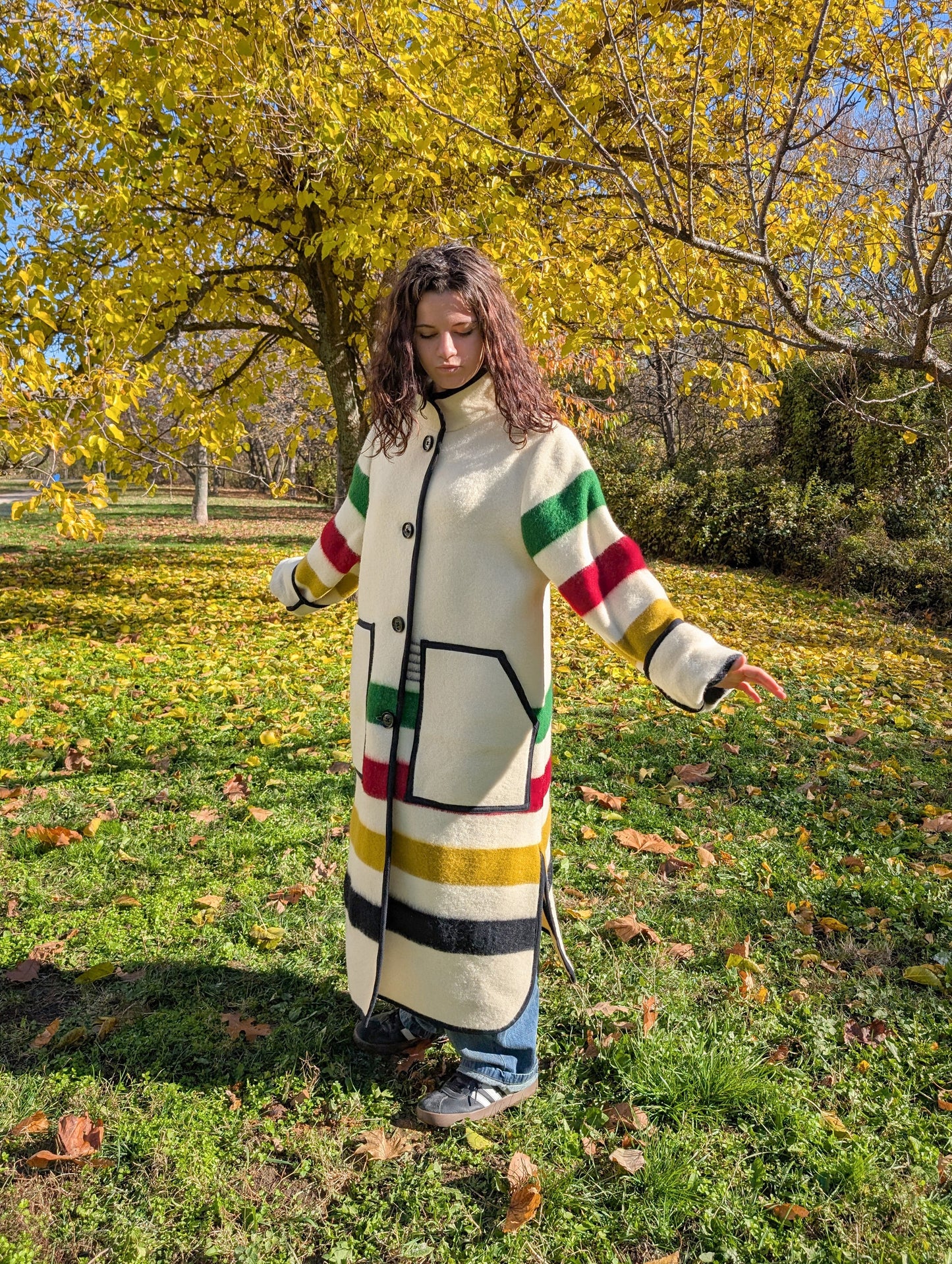 Person wearing a long colorful coat with cream base, duster coat, standing in a park with autumn leaves. The coat is single breasted with a stand collar. 