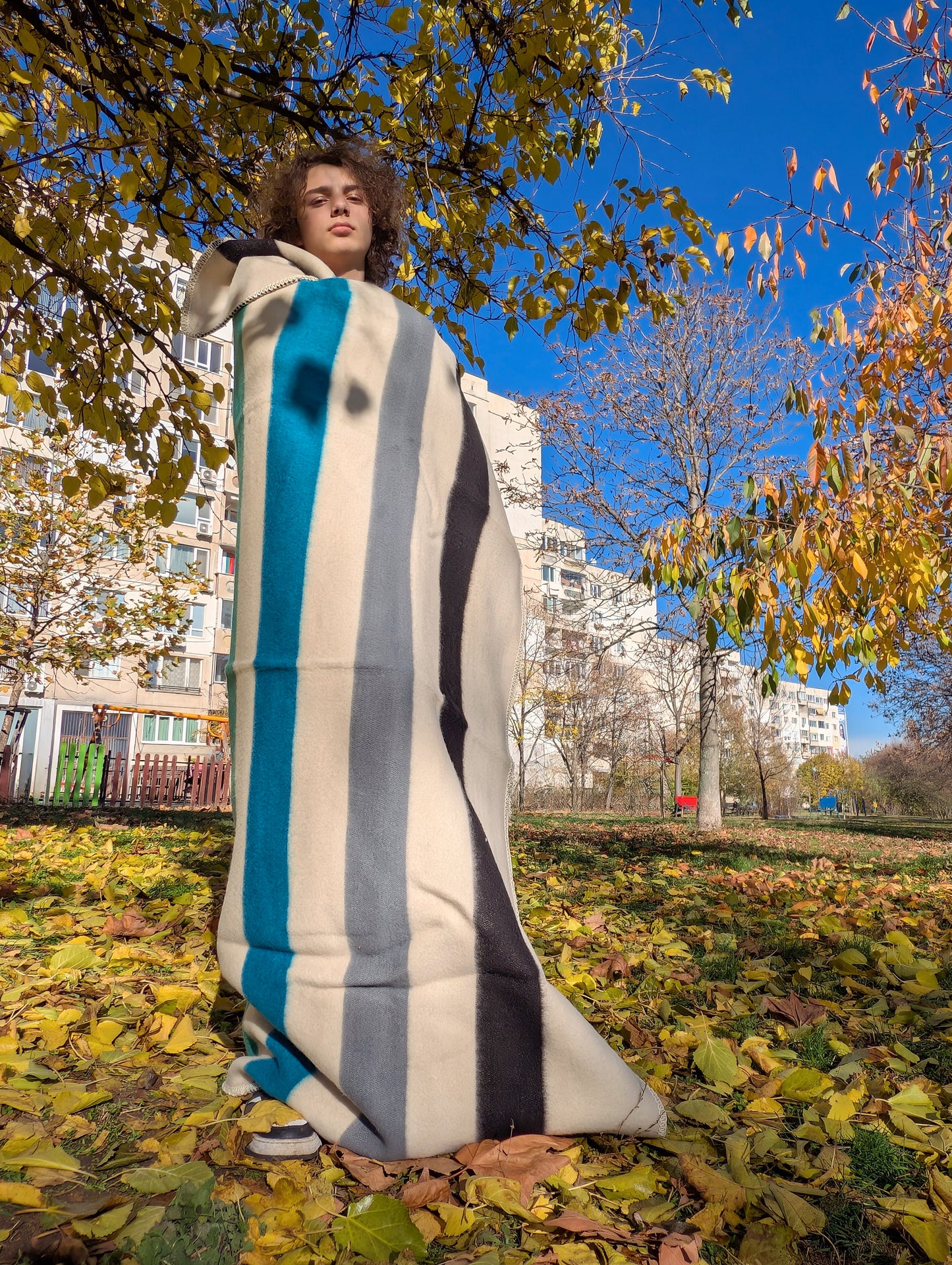 Person wrapped in a traditional multistriped blanket in an urban park with trees and buildings in the background.