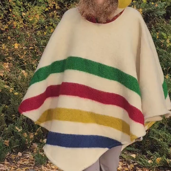 Video of woman with a hooded woolen poncho with 4 stripes in different colors on a light base. A unisex cape upcycled from a pure wool blanket.