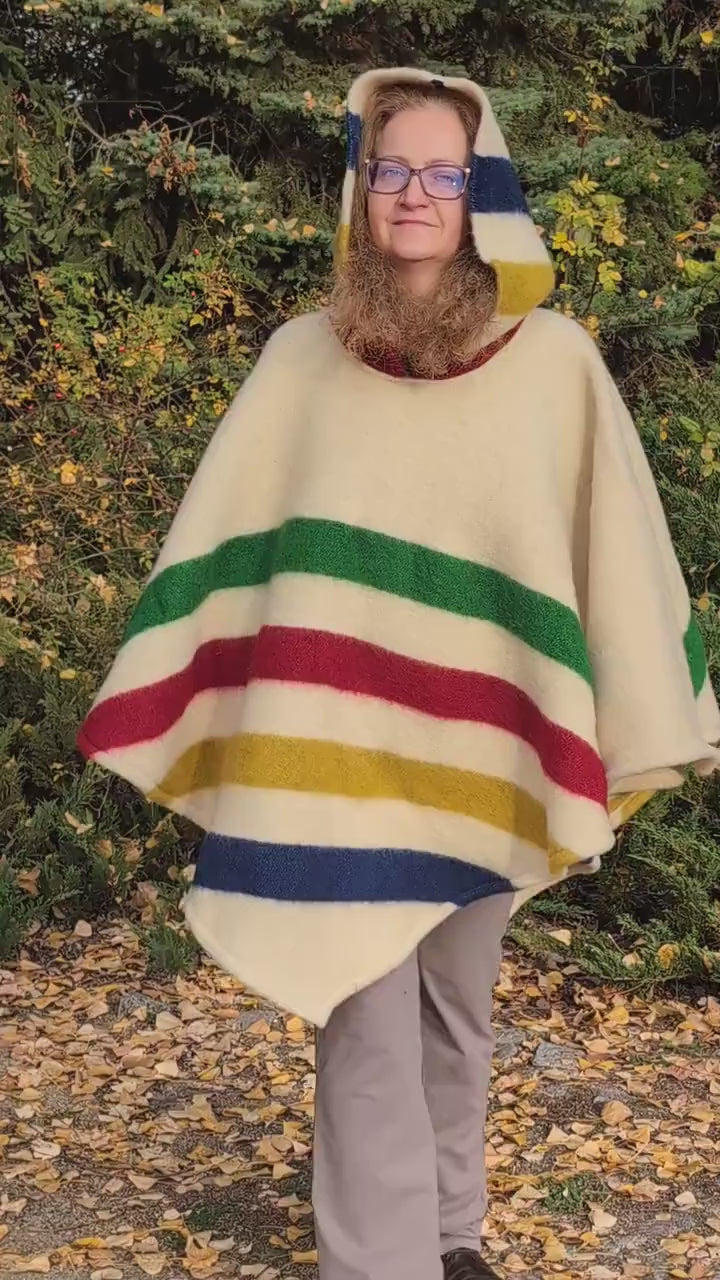 Video of woman with a hooded woolen poncho with 4 stripes in different colors on a light base. A unisex cape upcycled from a pure wool blanket.