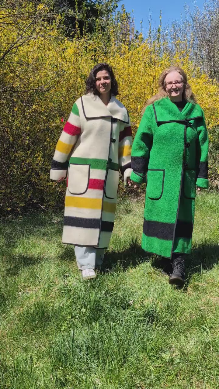 A video showing two women wearing same model long collar coats, one in solid traditional green with black stripe, and the other in ivory with multicolor stripes. Upcycled wool blanket dustercoats.